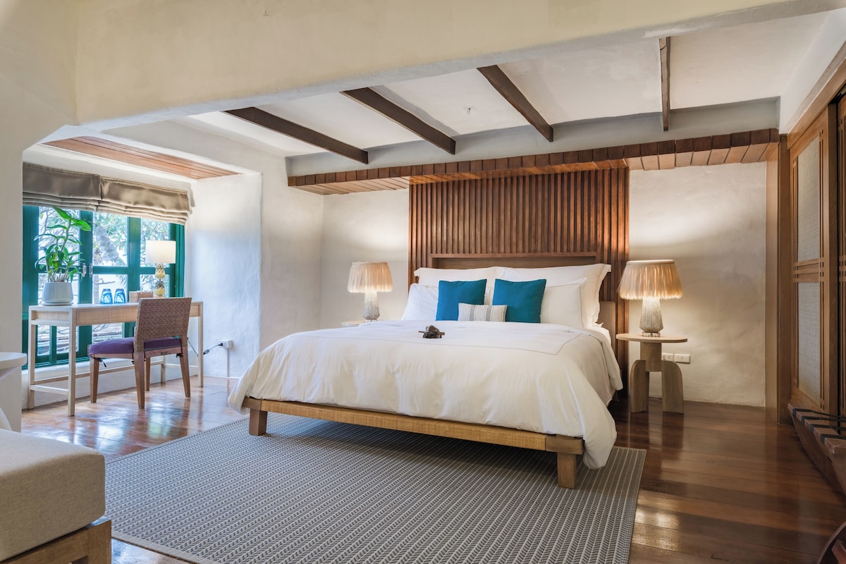 A bedroom with a large bed, wooden headboard, two bedside lamps, a desk with a chair, and a view of the outdoors.