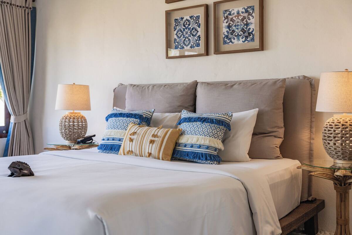 Una cama bien hecha con edredón blanco y varios almohadones, dos marcos de cuadros en la pared y dos lámparas de noche sobre mesitas de madera.