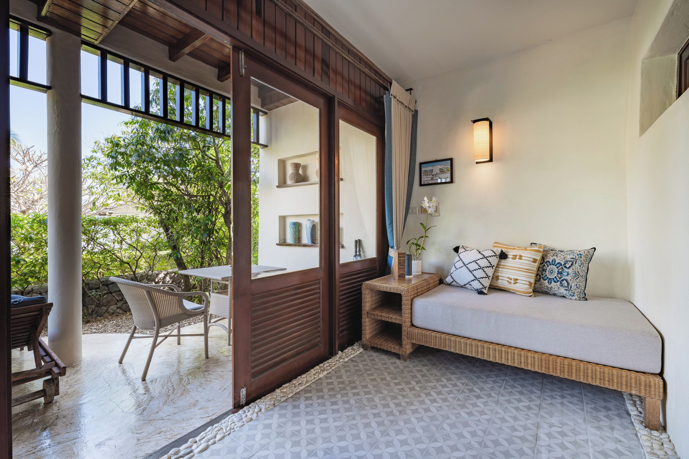 A cozy room with a wicker sofa, a small table, and a chair. There is a large window with a view of greenery outside.