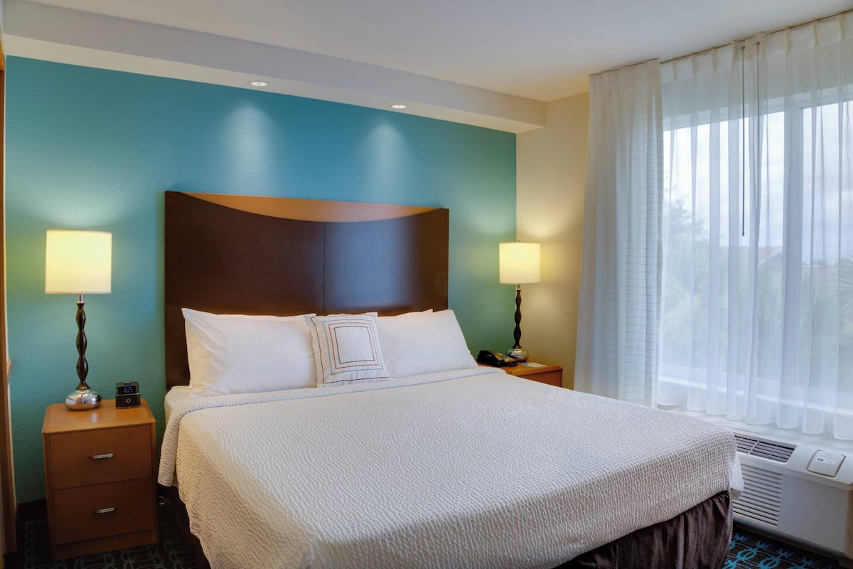 Habitación de hotel con una cama grande, dos mesitas de noche con lámparas, una mesita auxiliar y una ventana con cortinas traslúcidas.