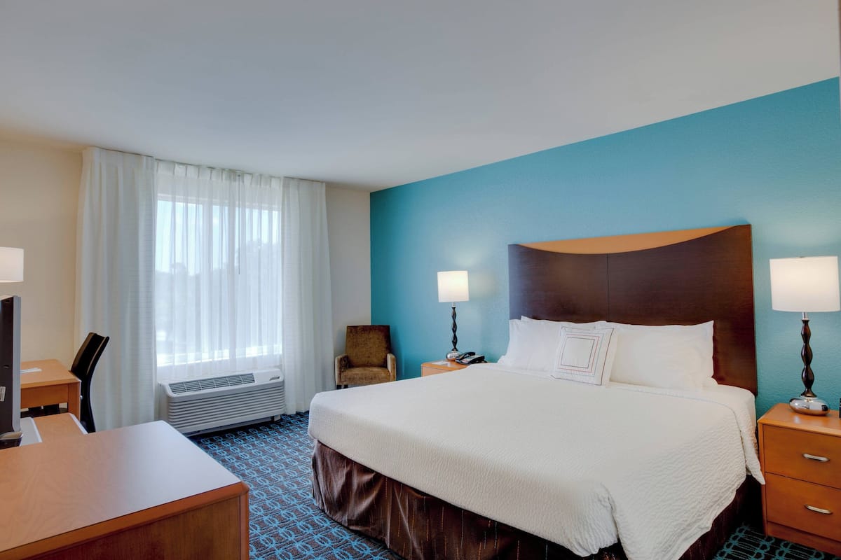 Una habitación de hotel con una cama grande, dos mesitas de noche con lámparas, un escritorio con una silla, una ventana con cortinas y un calentador de pared.