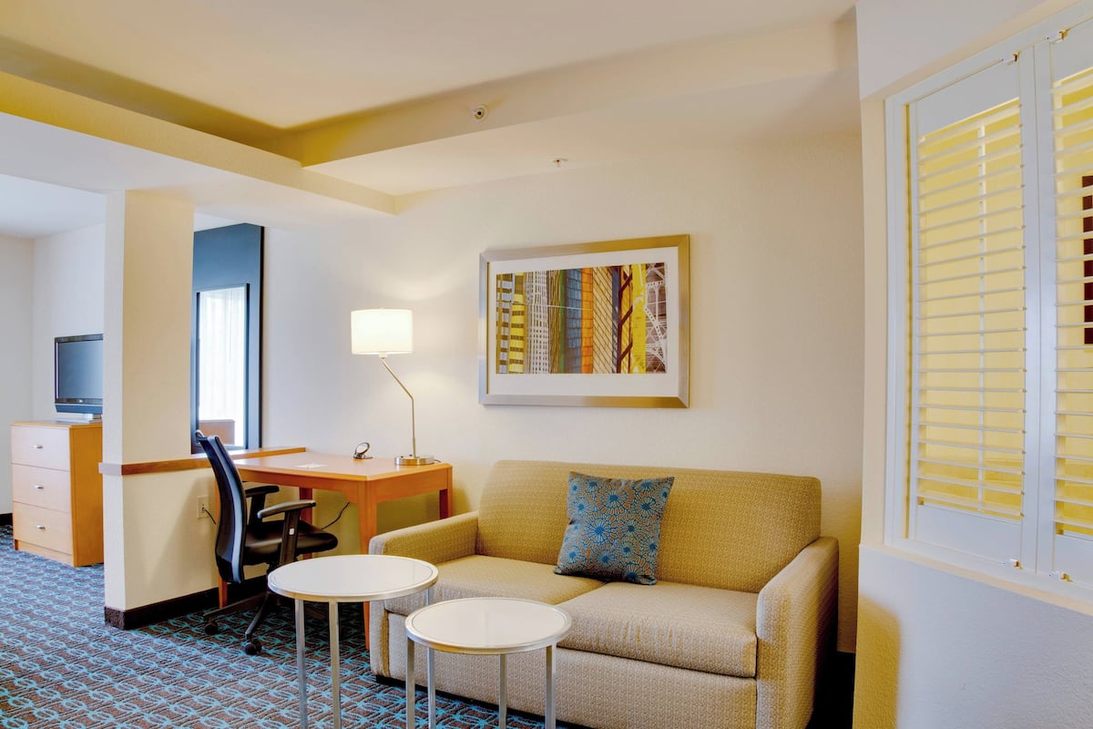 Habitación de hotel con sofá, dos mesitas, un escritorio, una silla, televisión y ventana con persianas.