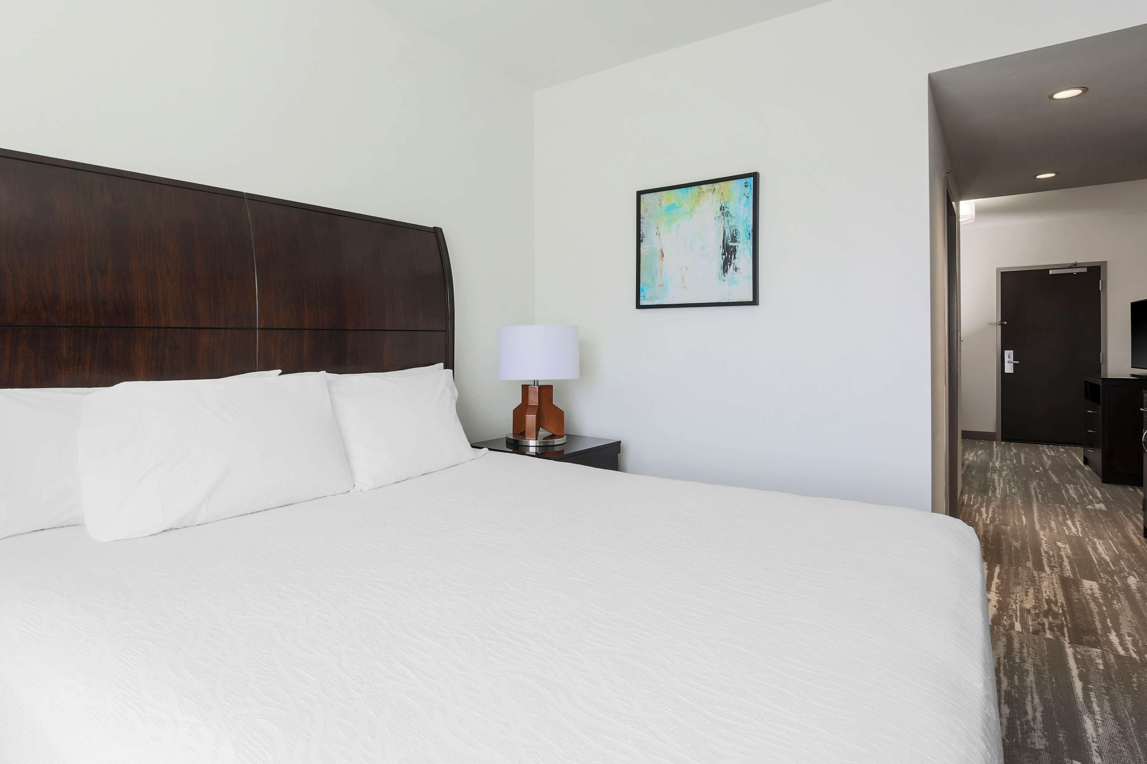 A hotel room with a large bed, a nightstand with a lamp, a framed picture on the wall, and a door leading to another room.