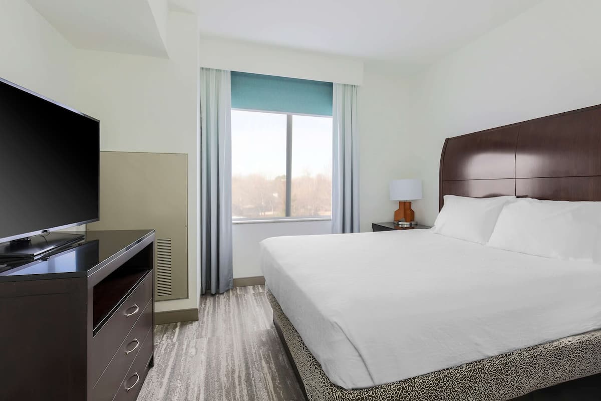 A hotel room with a large bed, a television on a dresser, a window with curtains, and a nightstand with a lamp.
