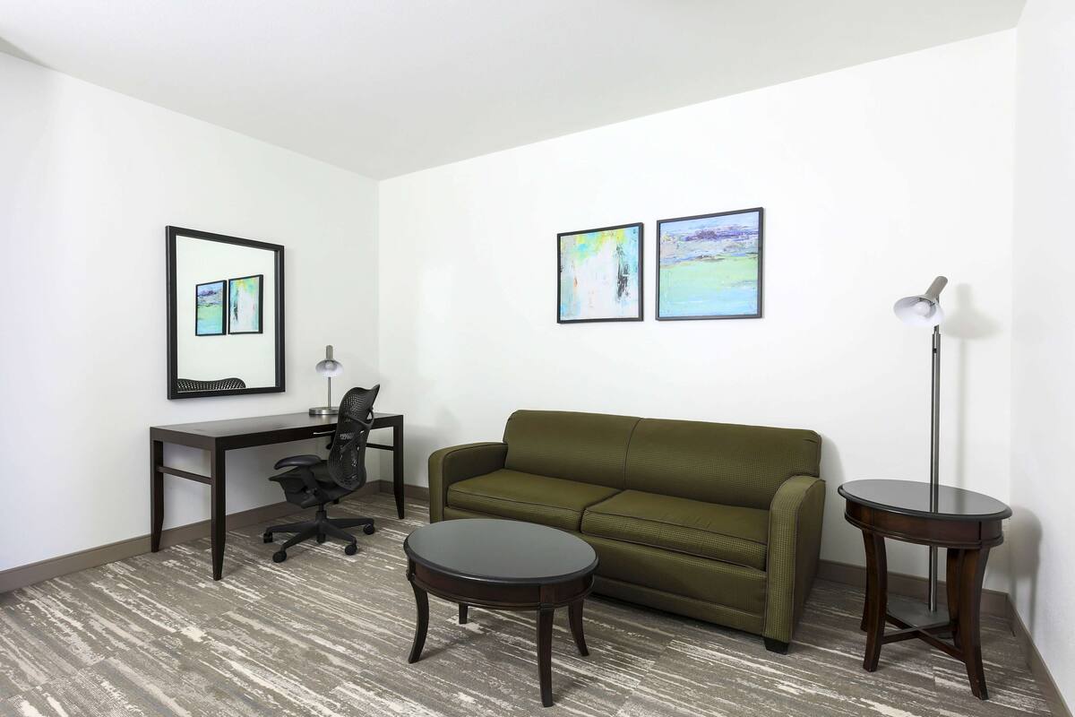 A room with a green sofa, a desk with a chair, a round table, and two framed artworks on the wall.
