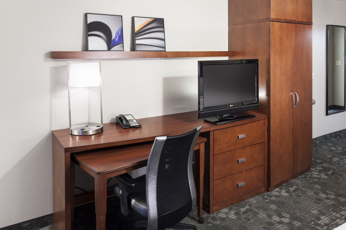 A hotel room with a wooden desk, a black office chair, a flat-screen TV, a telephone, and a cabinet.