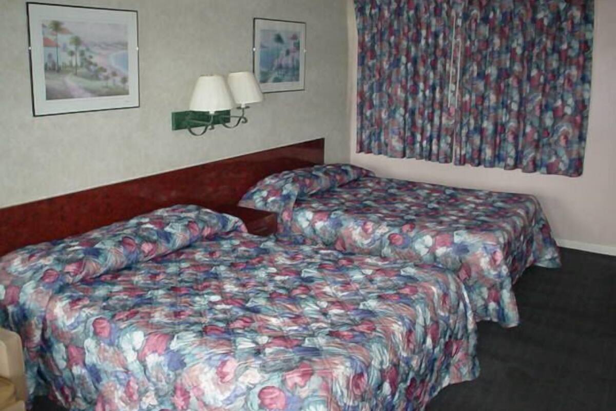 Habitación de hotel con dos camas, cada una con ropa de cama floral, cabecero, mesita de noche, dos lámparas de pared y una ventana con cortinas estampadas.