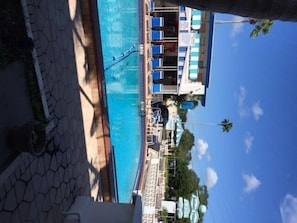 Zona de piscina con sillas de descanso y sombrillas, un cielo azul despejado y un cartel que dice 'Bellas Vistas'.