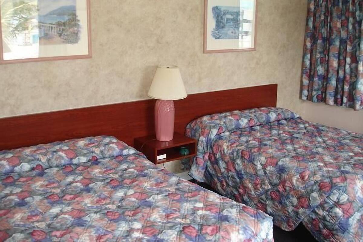 Habitación de hotel con dos camas, una mesita de noche con lámpara y dos cuadros en la pared.