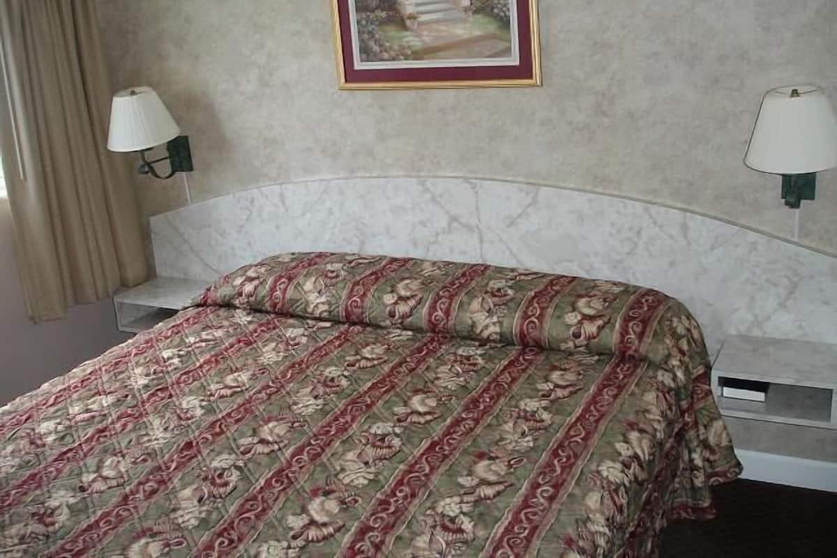 Una cama bien hecha con una colcha estampada, dos lámparas de noche, un cuadro enmarcado en la pared y una ventana con cortinas.