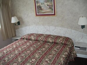 A neatly made bed with a floral bedspread, two bedside lamps, a framed picture on the wall, and a window with curtains. - Belleair Village Motel (Largo)