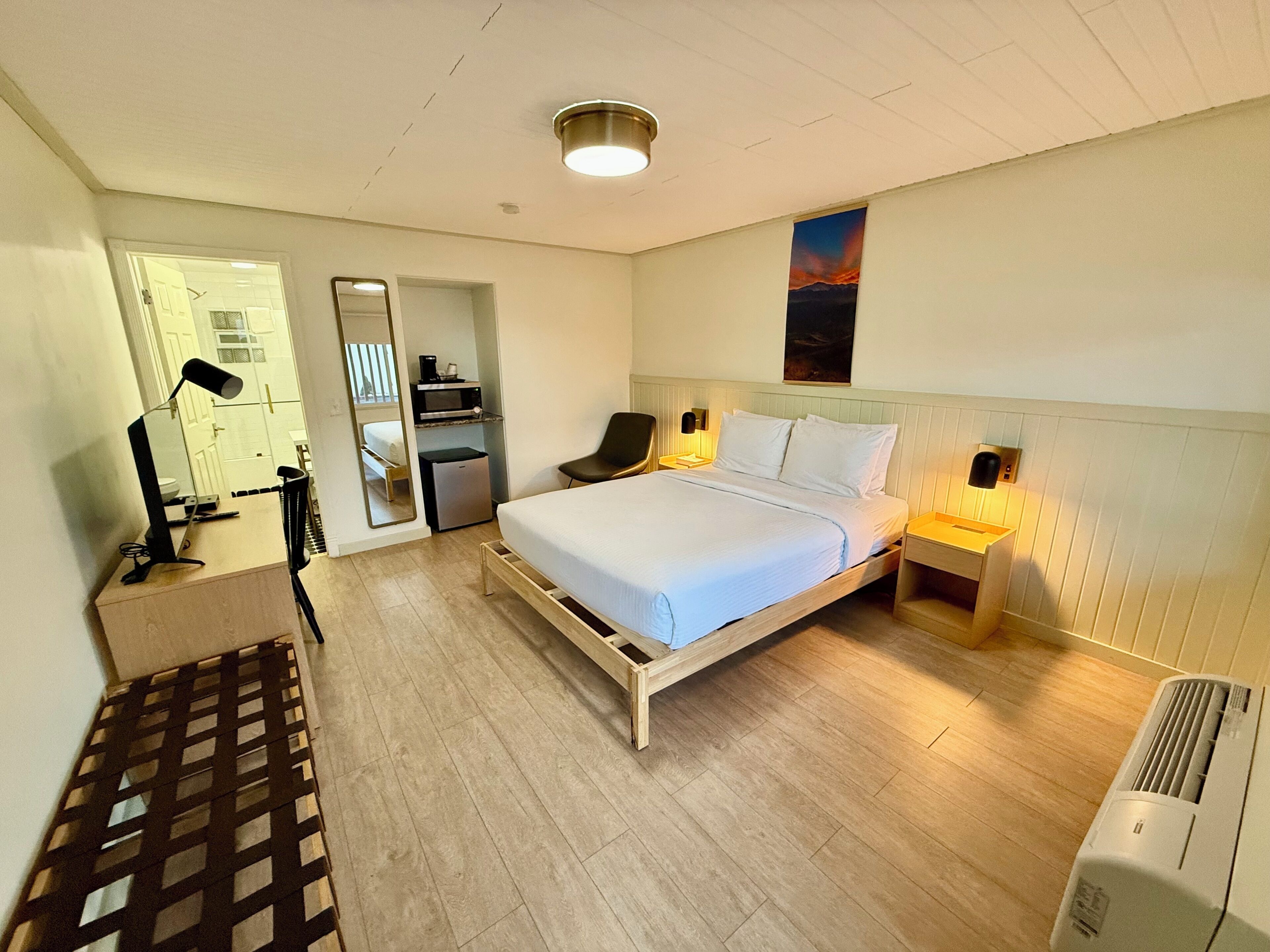 Quarto de hotel com cama, mesas de cabeceira, escrivaninha com luminária, espelho, armário e aquecedor.