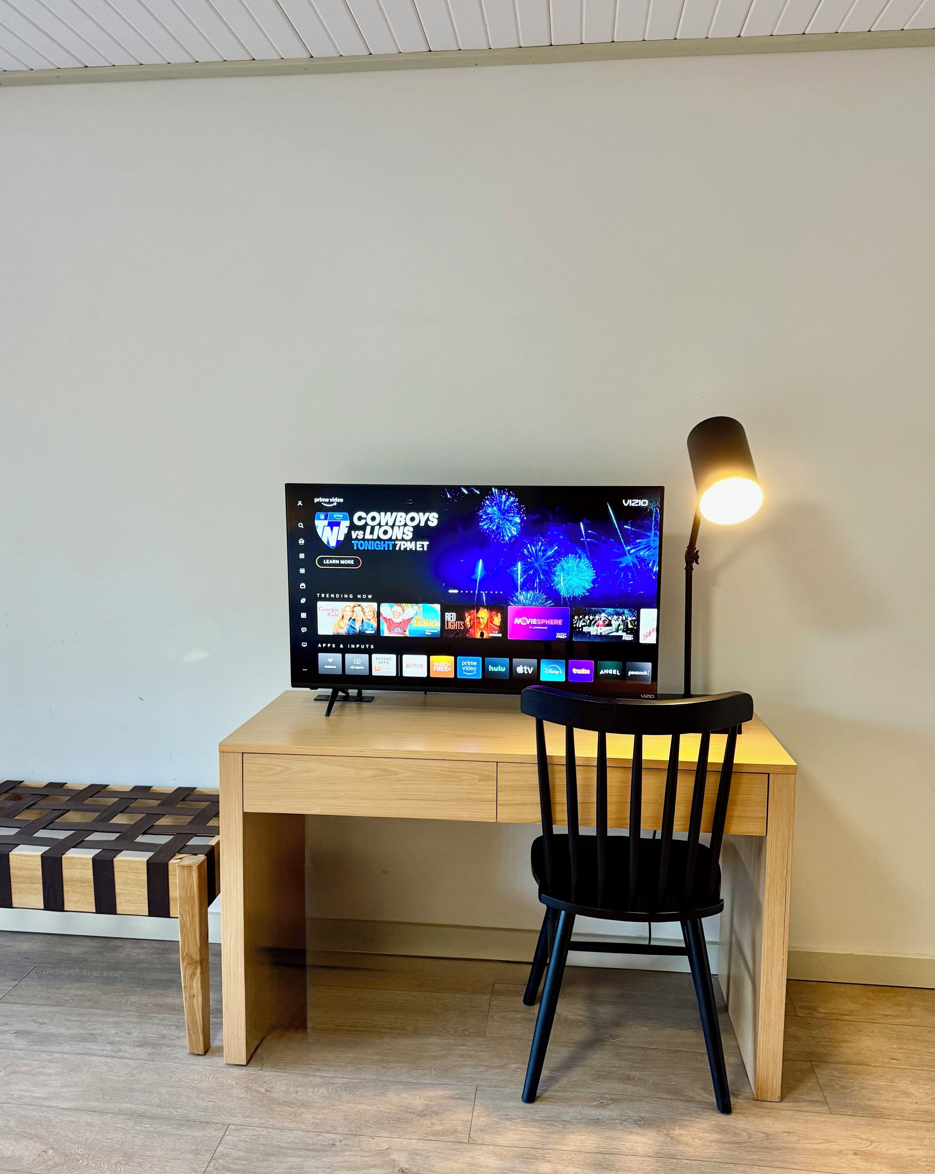 Un escritorio de madera con una televisión de pantalla plana mostrando un evento deportivo, una silla negra y una lámpara de escritorio.
