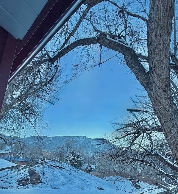 A snow-covered landscape with a clear sky, a tree in the foreground, and a distant view of a house and mountains.
