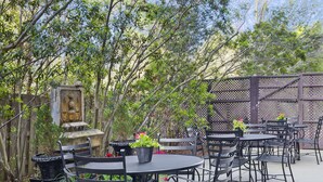 Un espace extérieur aménagé en terrasse, avec des tables rondes et des chaises en métal, entouré d’arbres et d’une clôture décorative.