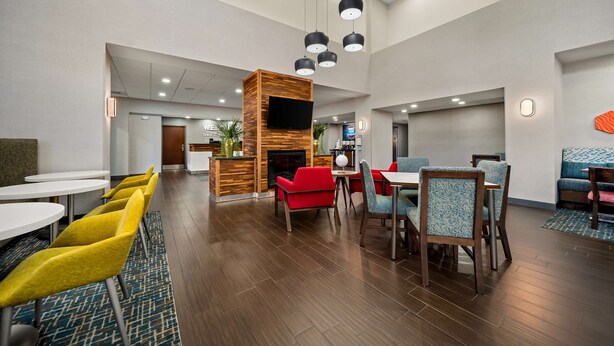 A spacious indoor area with multiple seating arrangements, including yellow chairs, red sofas, and blue chairs. There is a wooden partition with a flat-screen TV and a fireplace. The area is well-lit with ceiling lights and has a patterned carpet. - Hampton Inn Suites Opelika I85 Auburn Area (Opelika)