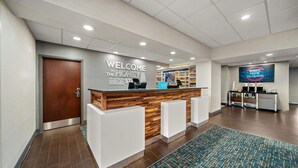 A reception area with a wooden reception desk, a 'Welcome to the Honey Bee Stay' sign, a digital screen displaying 'Home Alabama', and a blue patterned rug. - Hampton Inn Suites Opelika I85 Auburn Area (Opelika)