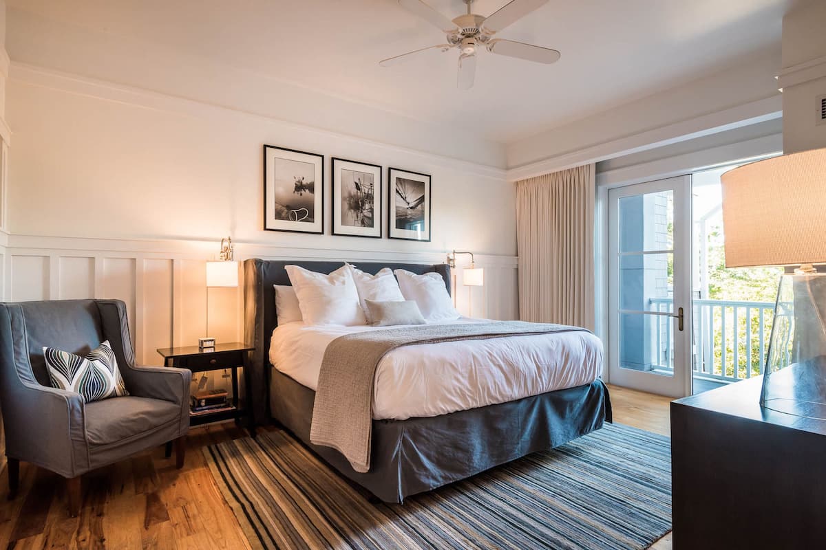 A hotel room with a large bed, a chair, a desk, and a balcony with curtains.