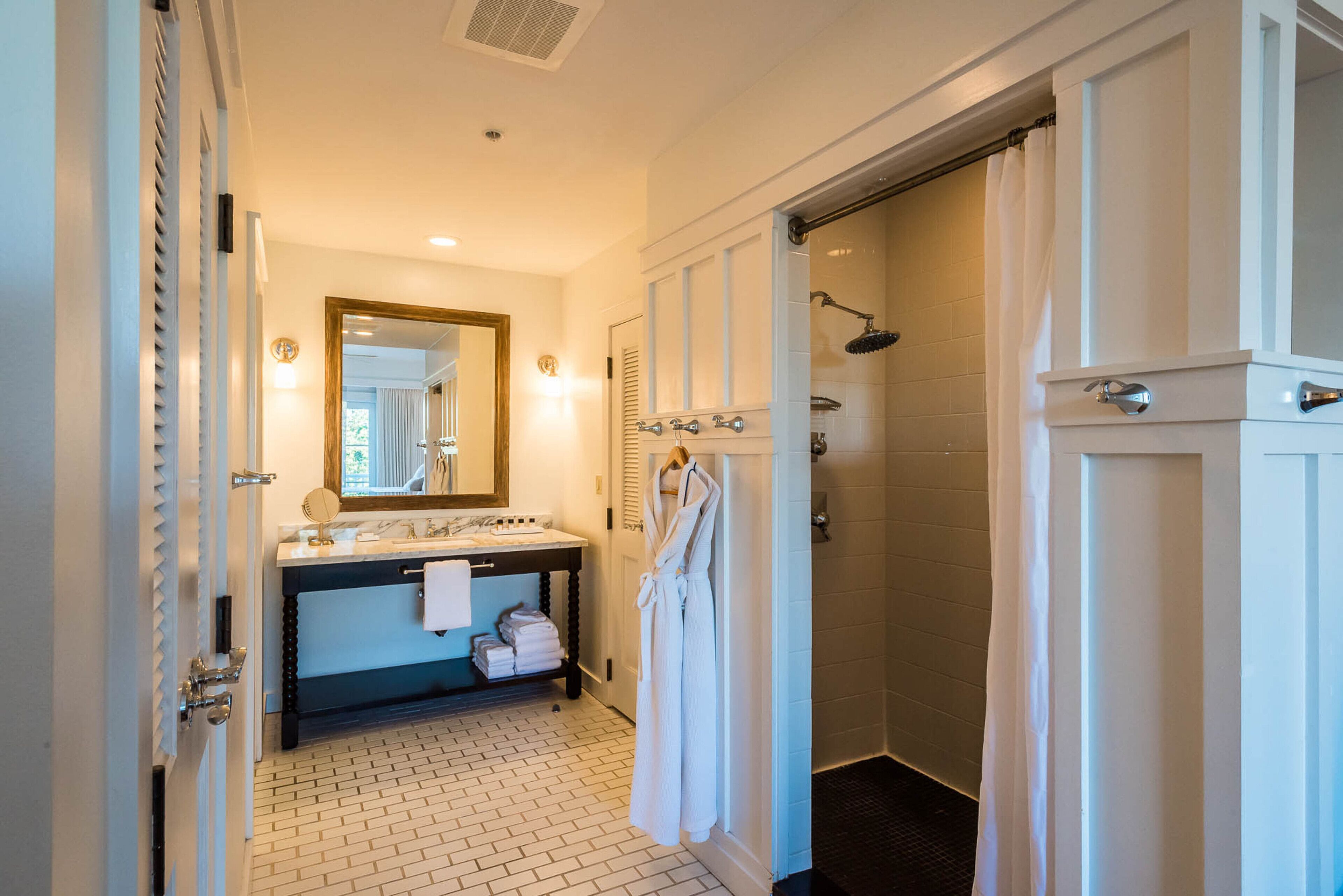 Une salle de bain avec un sol carrelé de carreaux blancs, un lavabo avec un miroir, un peignoir suspendu à un porte-serviettes et un espace douche.