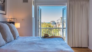 A bedroom with a bed, a nightstand, a lamp, a window with curtains, and a view of buildings outside. - WaterSound Inn (Santa Rosa Beach)
