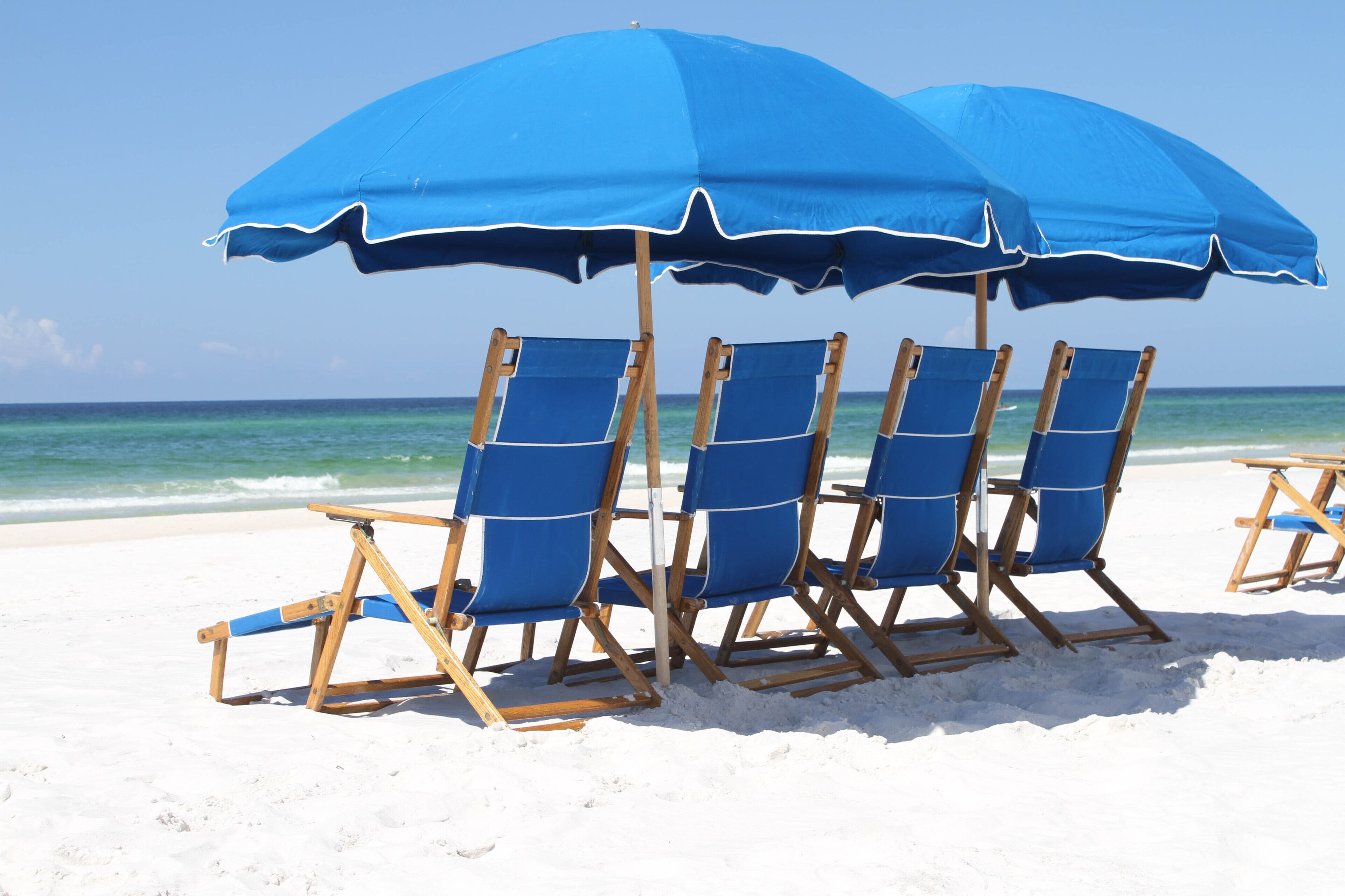Four blue beach chairs under a large blue umbrella on a sandy beach with the ocean in the background.