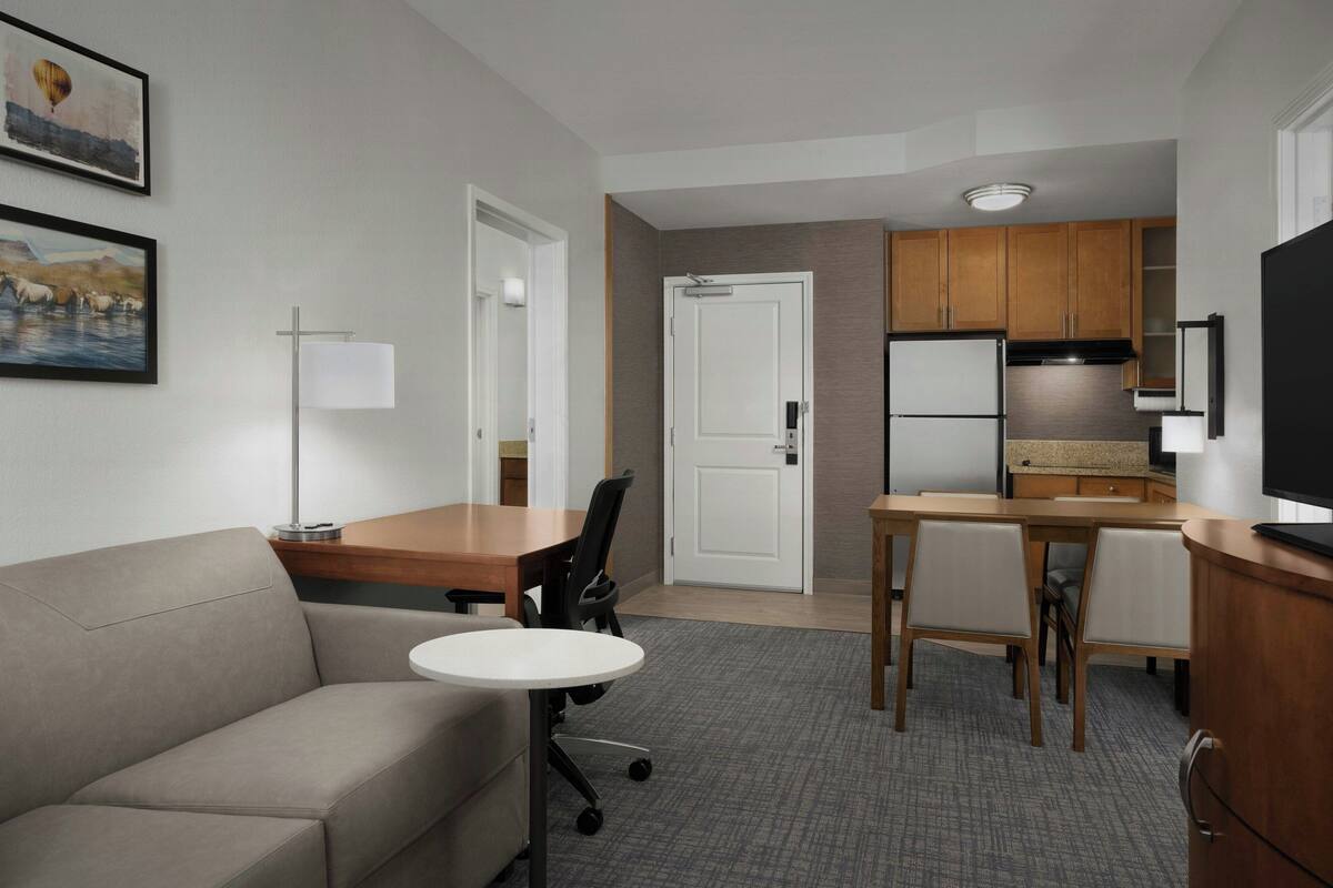 A hotel room with a sofa, a small round table, a desk with chairs, a kitchenette, and a TV.