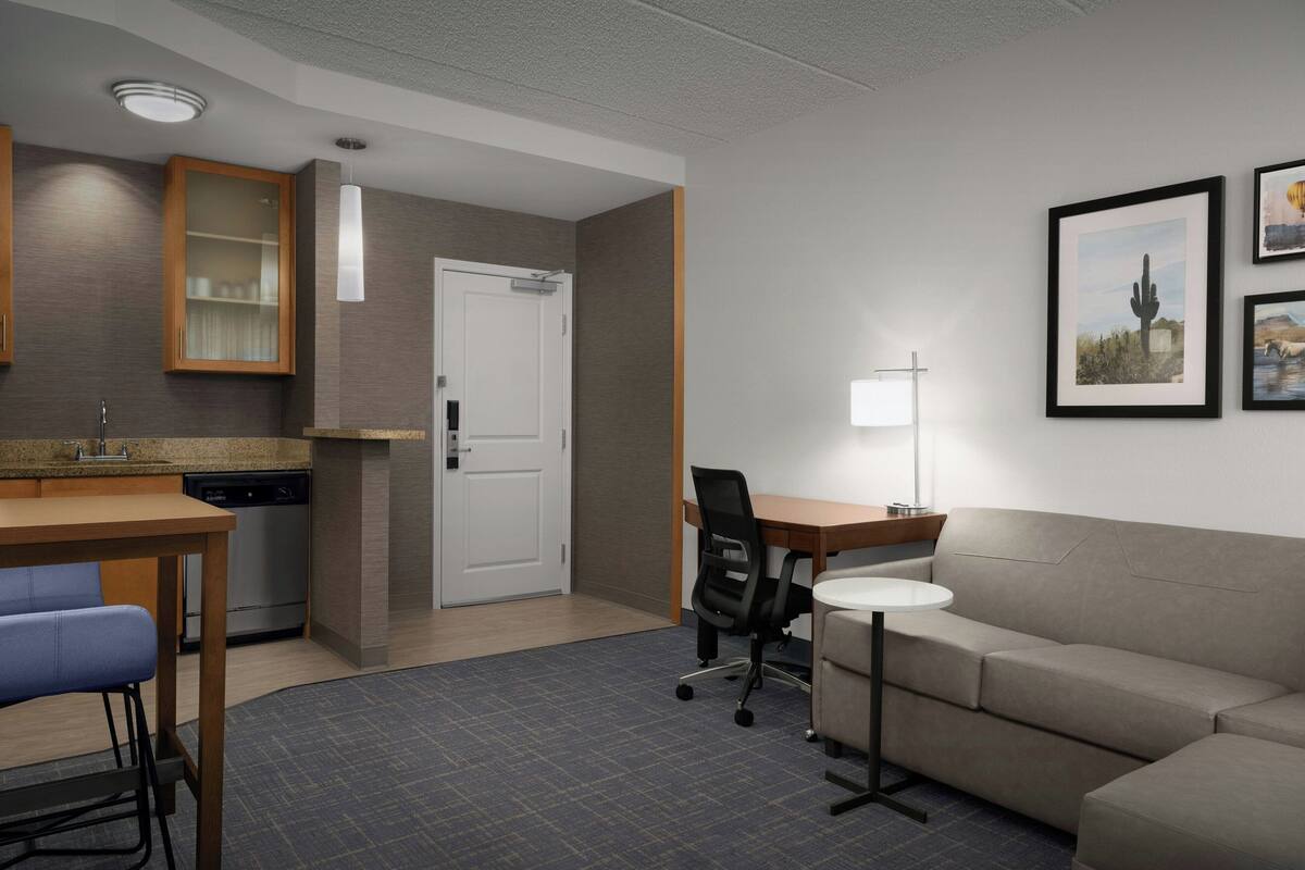 A modern hotel room with a kitchenette, a dining area, a sofa, and a desk.