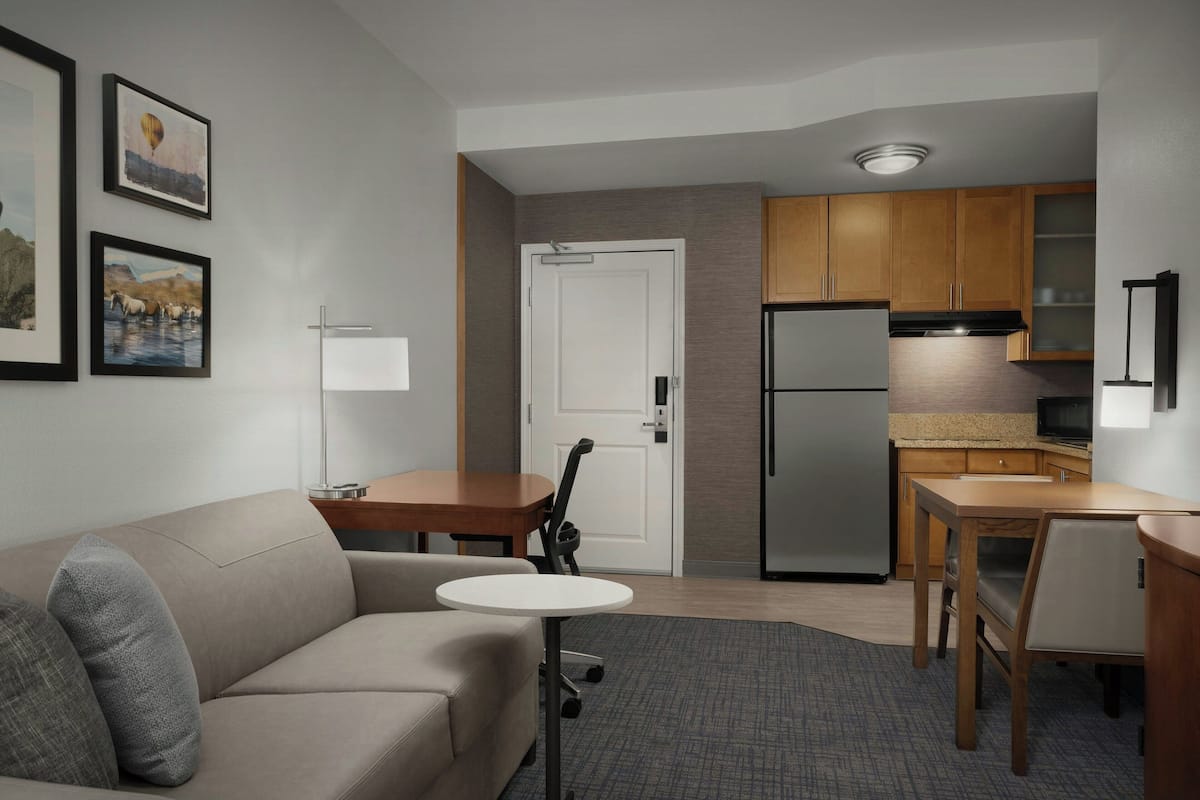 A compact living space with a sofa, a small round table, a kitchenette with wooden cabinets, a refrigerator, and a door.