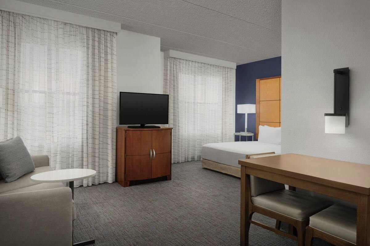 A hotel room with a bed, a TV on a wooden cabinet, a small round table, a sofa, and a desk with two chairs.