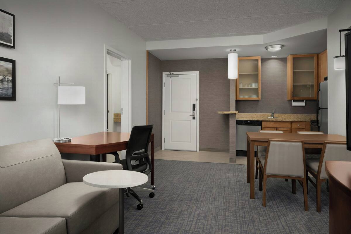 A compact hotel room with a kitchenette, a dining area with a table and chairs, a sofa, and a small round table.