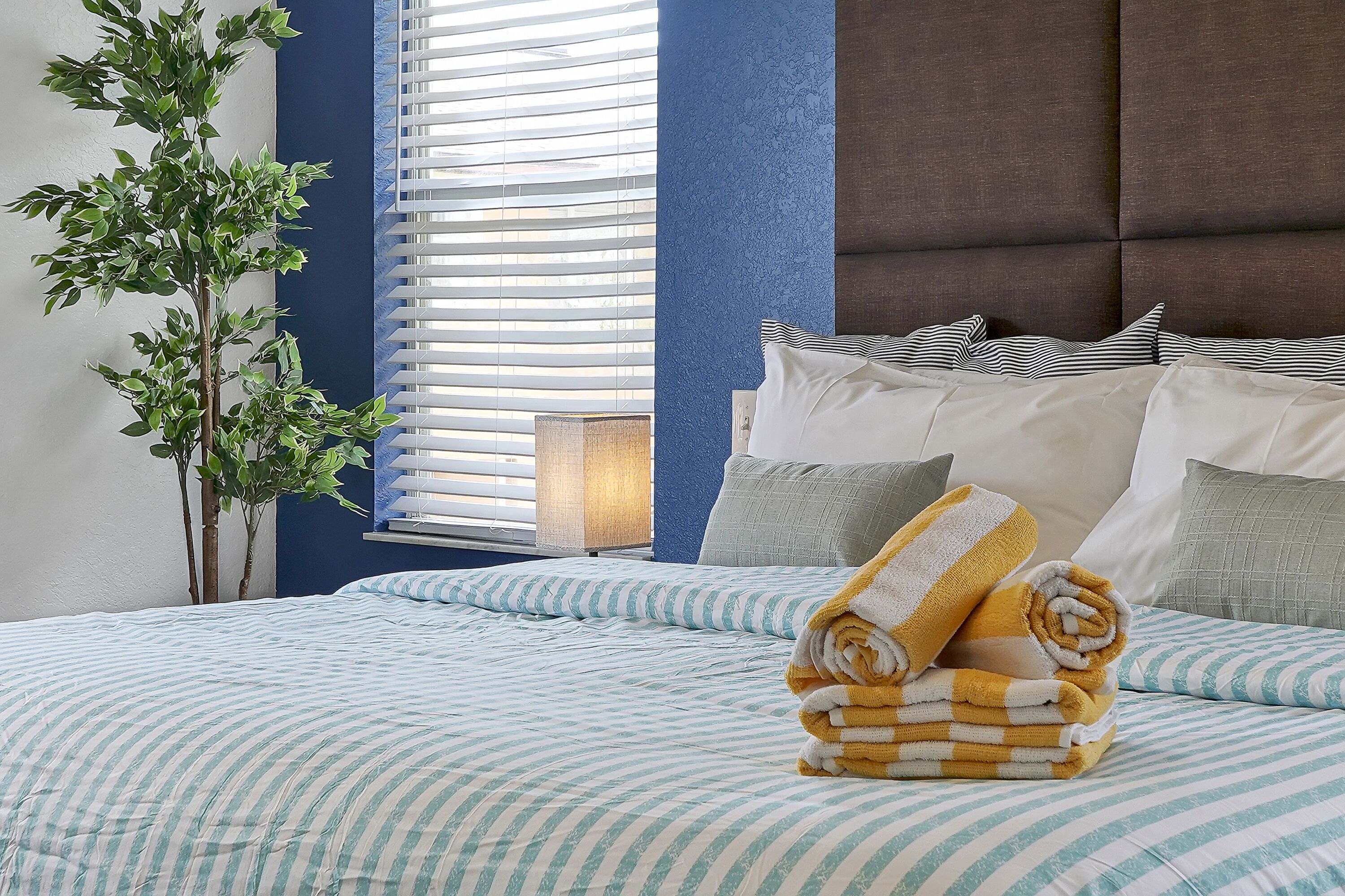 A neatly made bed with striped bedding, a potted plant, and a window with blinds.