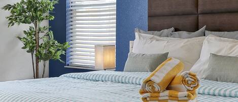A neatly made bed with striped bedding, a potted plant, and a window with blinds.