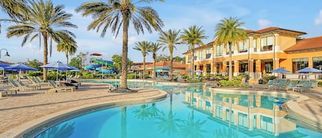 Un espace piscine aménagé dans un complexe hôtelier, avec des palmiers, des chaises longues et un bâtiment doté d’un toit en tuiles.