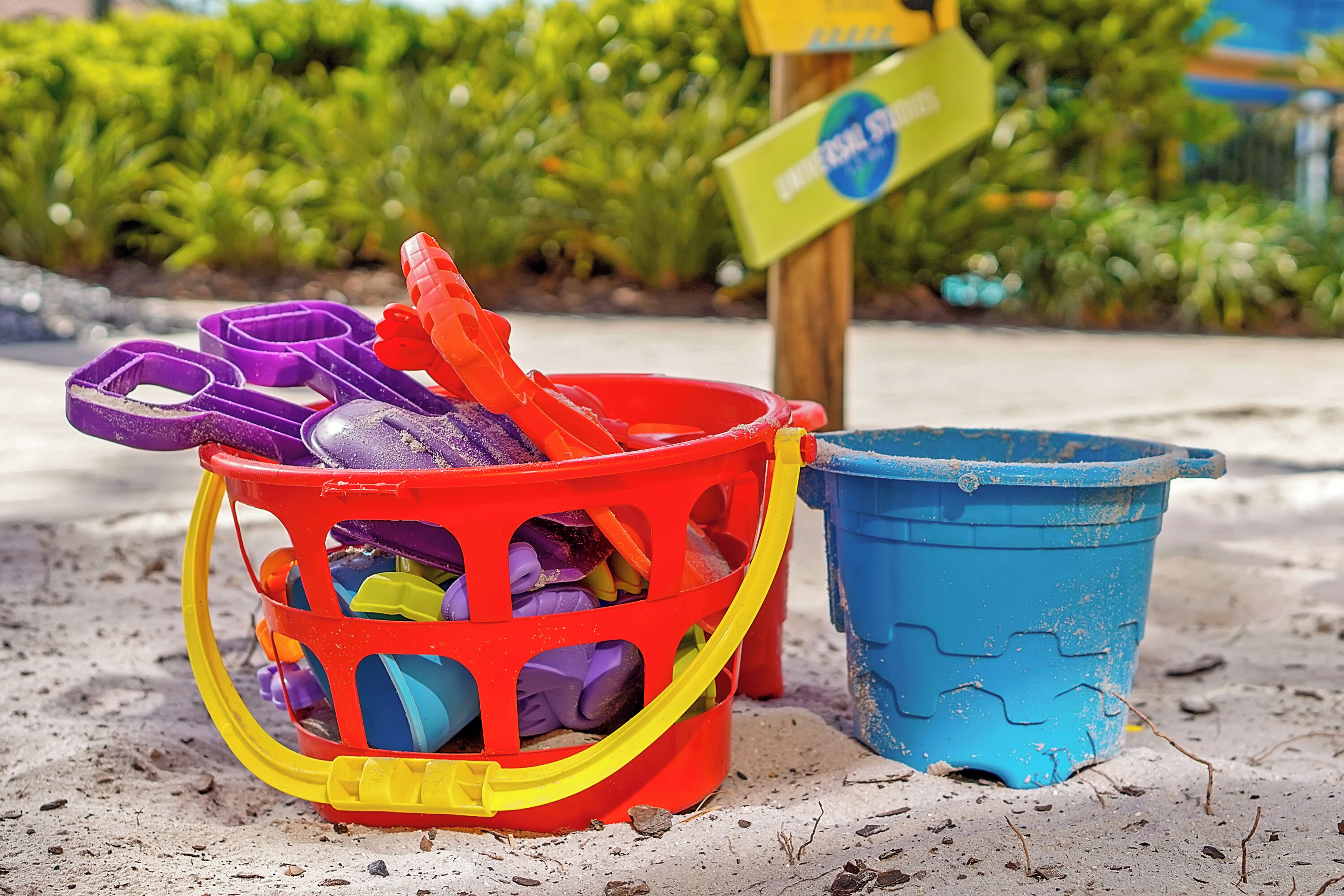 A beach scene with a red and yellow plastic bucket filled with colorful beach toys, a blue plastic bucket, and a sign in the background.