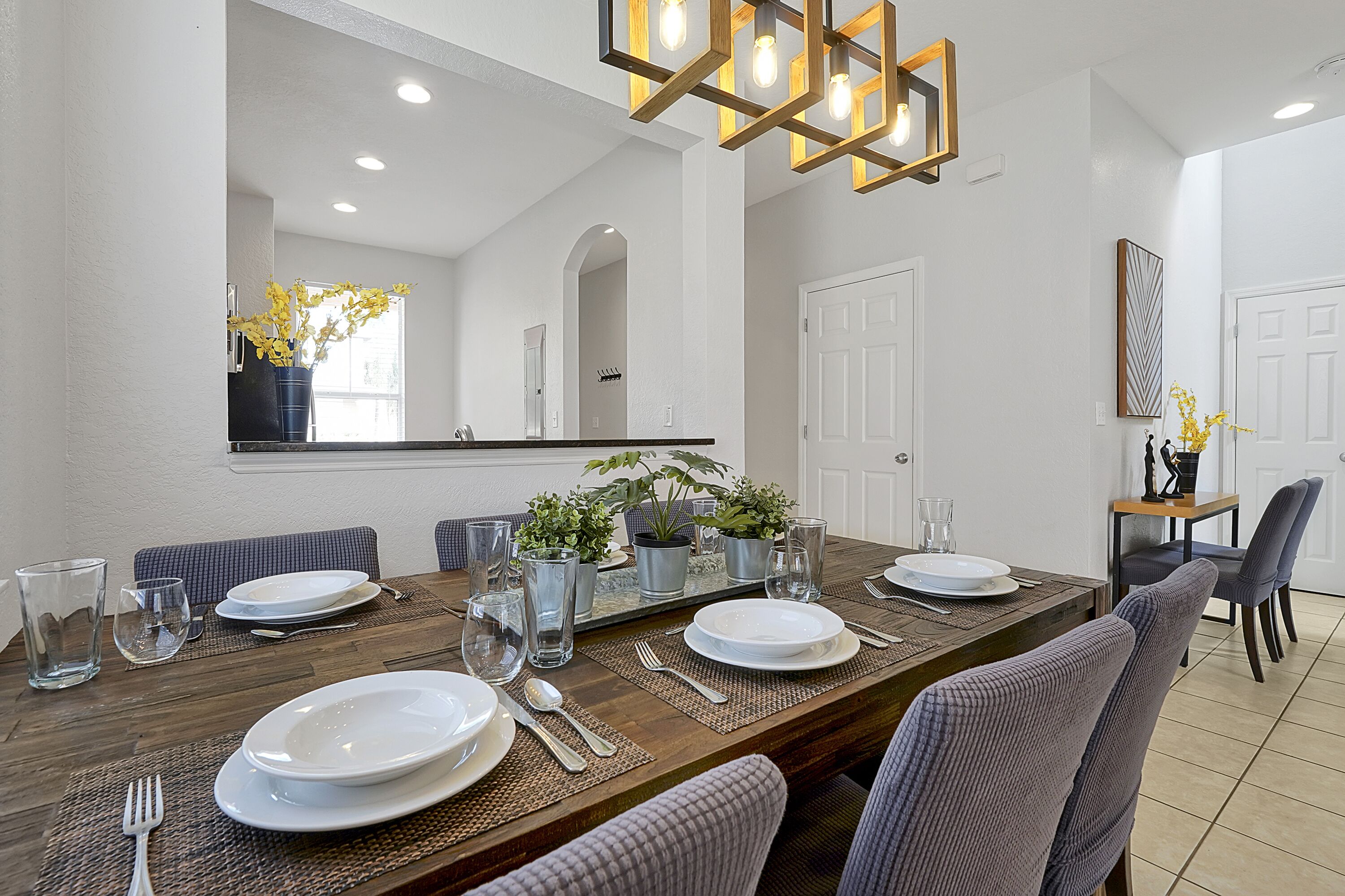 A dining area with a wooden table set for four, surrounded by chairs, with a large mirror above and a potted plant on the table.