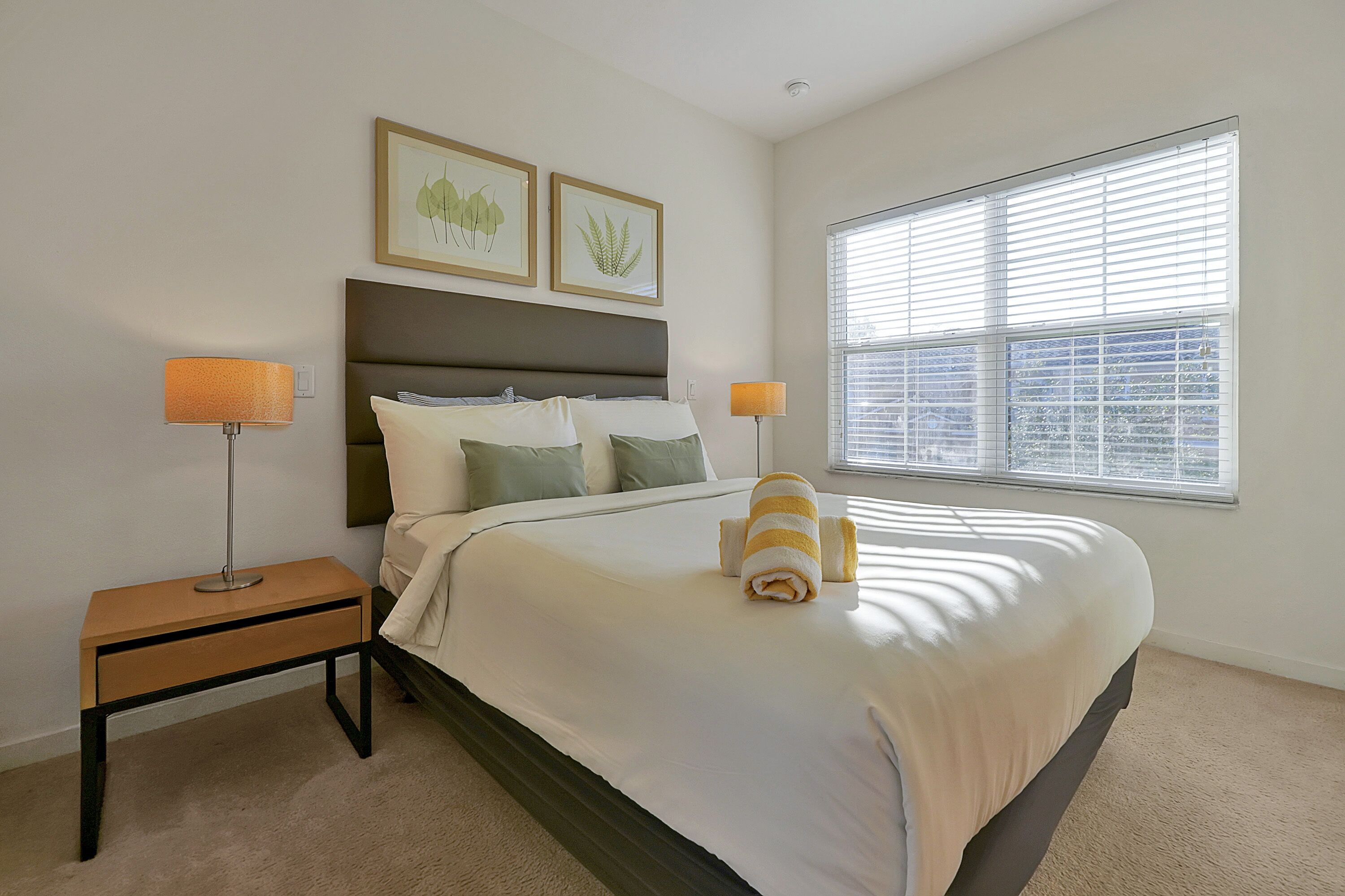 A neatly made bed with a white comforter and two pillows, a bedside table with a lamp, and a window with blinds.