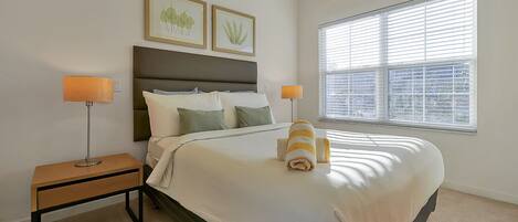A neatly made bed with a white comforter and two pillows, a bedside table with a lamp, and a window with blinds.