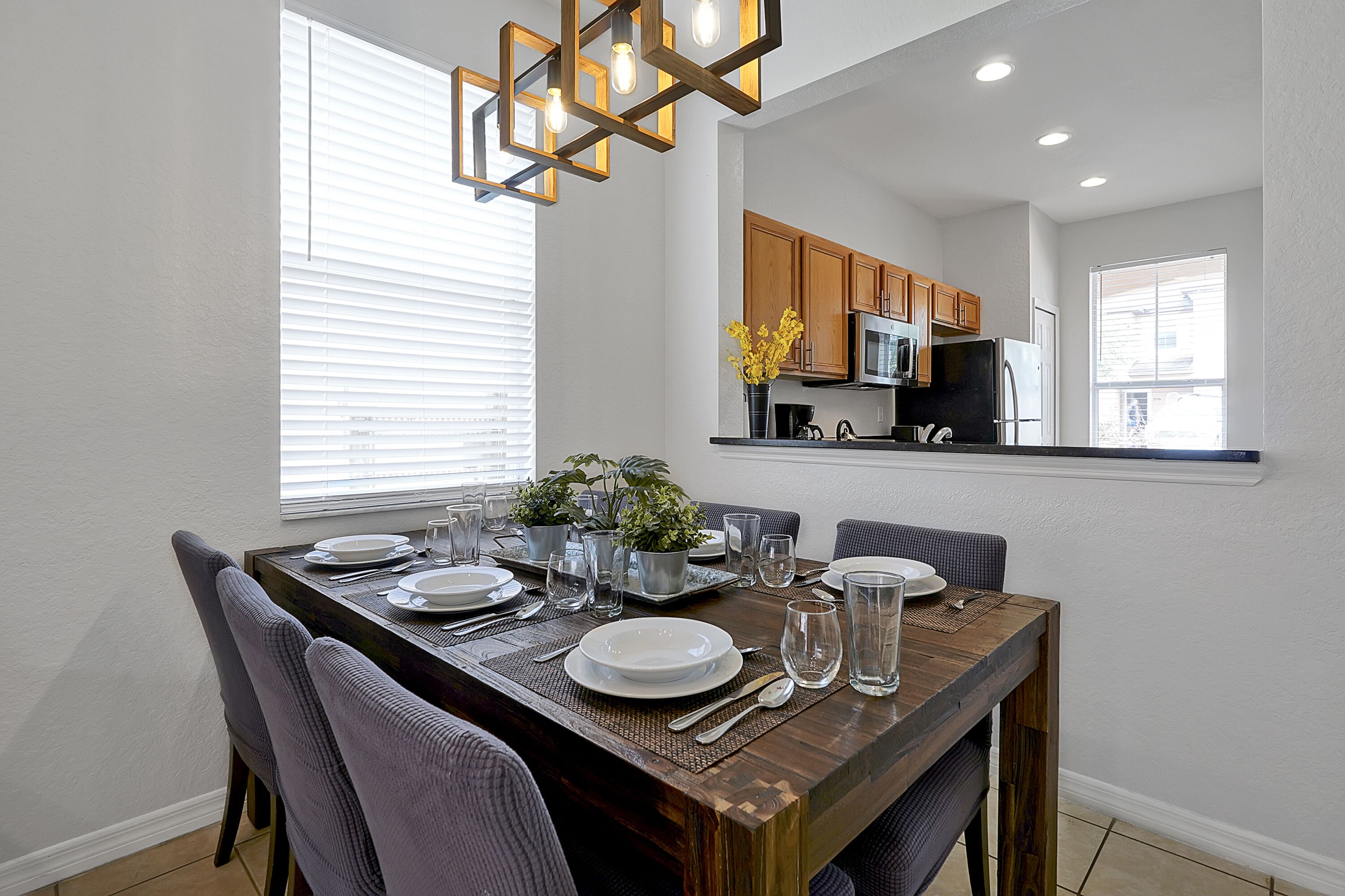 Un comedor con una mesa de madera dispuesta para cuatro personas, rodeada de sillas. Se observa una cocina al fondo con armarios de madera y electrodomésticos modernos.