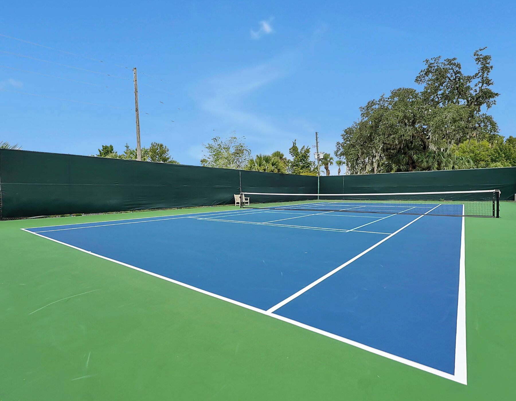 Una cancha de tenis con red, rodeada por una cerca verde y árboles.