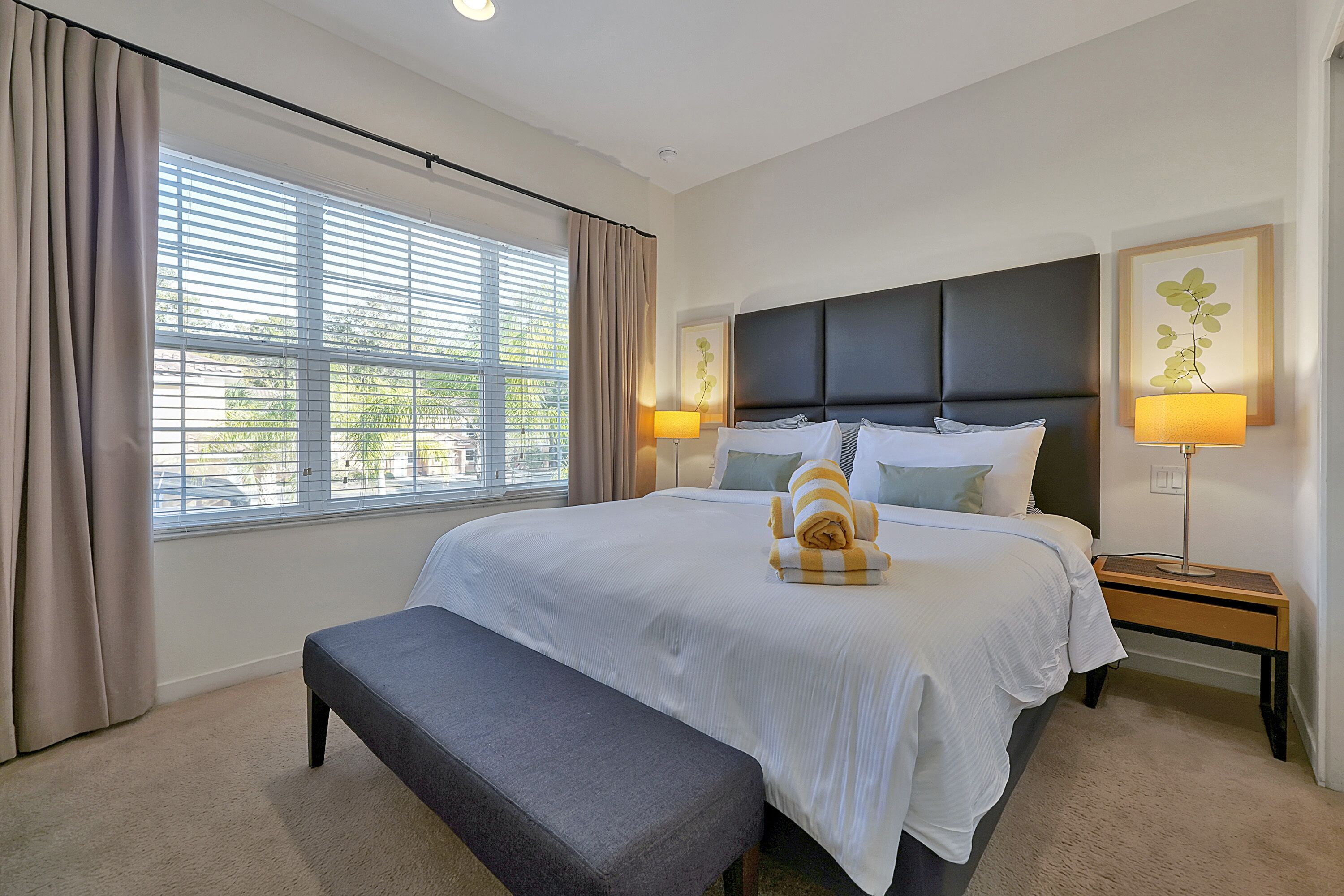 A neatly arranged bedroom with a large bed, a bench, two bedside tables with lamps, and a window with curtains.