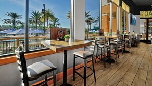 A restaurant with a view of palm trees and a pool. It features wooden tables and chairs, and large windows.