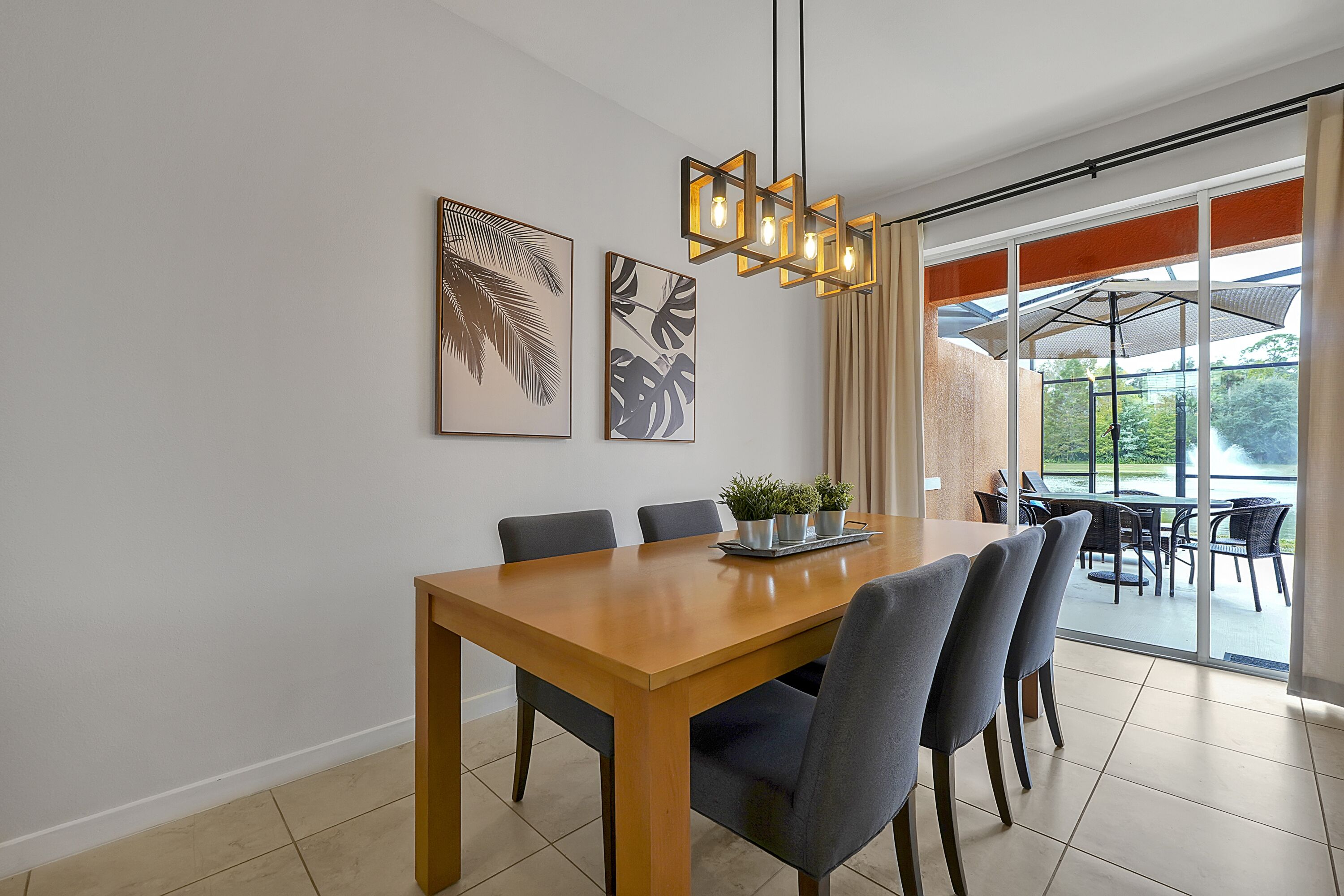 A dining area with a wooden table, six chairs, and a hanging light fixture. There are two framed wall art pieces and a potted plant on the table.