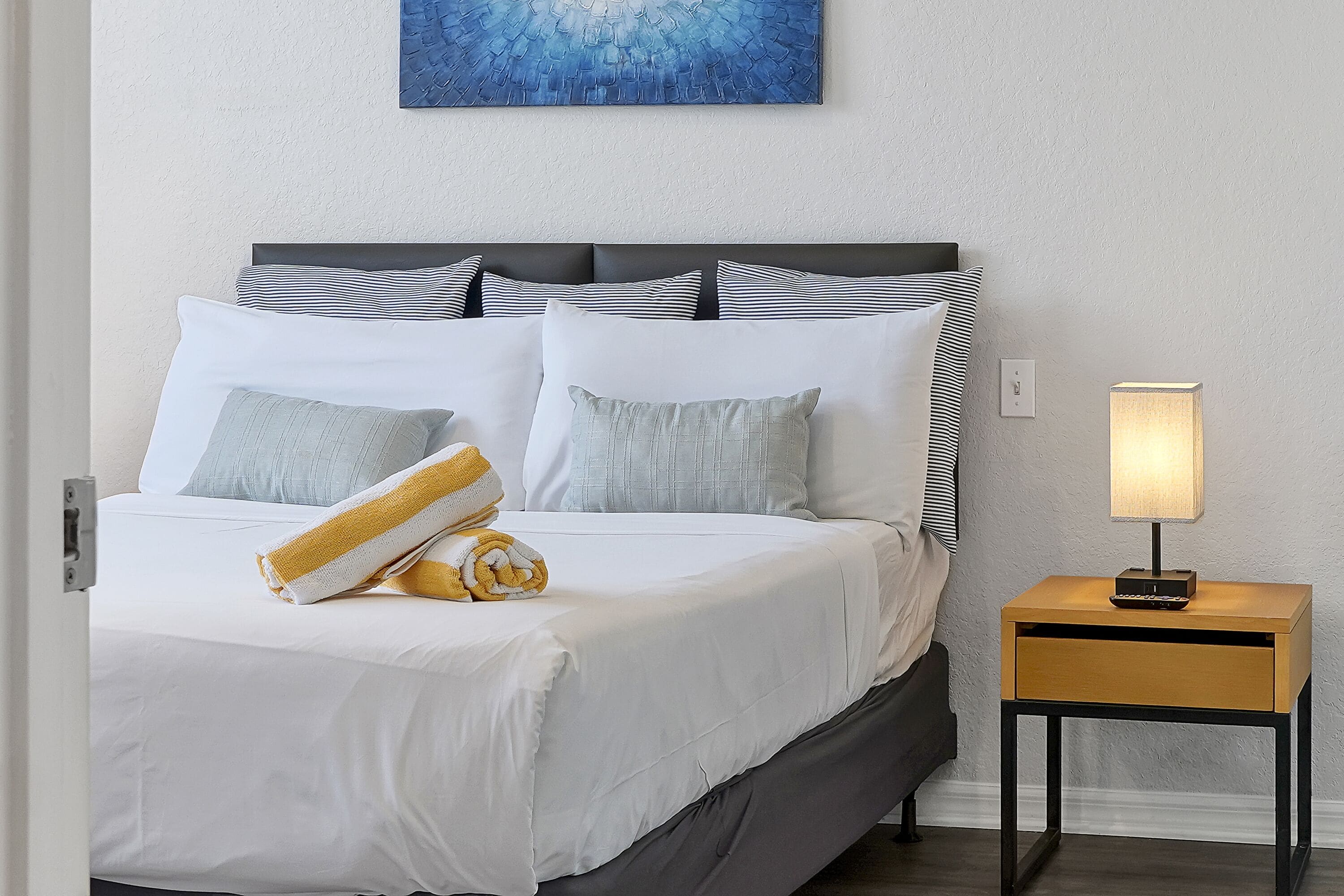 Ein ordentlich bezogenes Bett mit weißen Leinen und gestreiften Kissen, ein Nachttisch aus Holz mit einer Lampe und ein blaues abstraktes Gemälde an der Wand.
