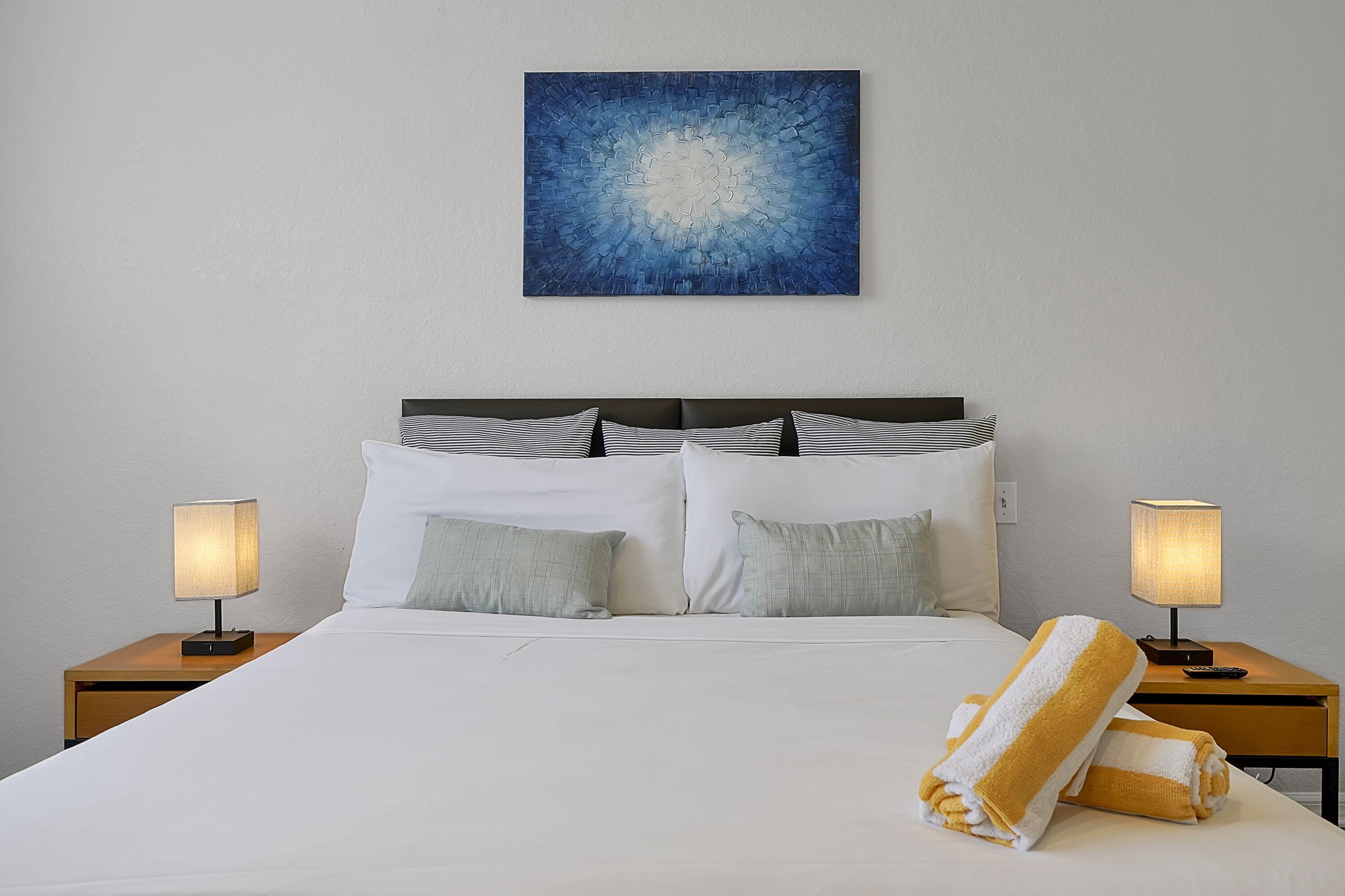 Una cama bien hecha con sábanas blancas y dos almohadas, flanqueada por dos mesitas de noche con lámparas, y un cuadro abstracto azul en la pared.
