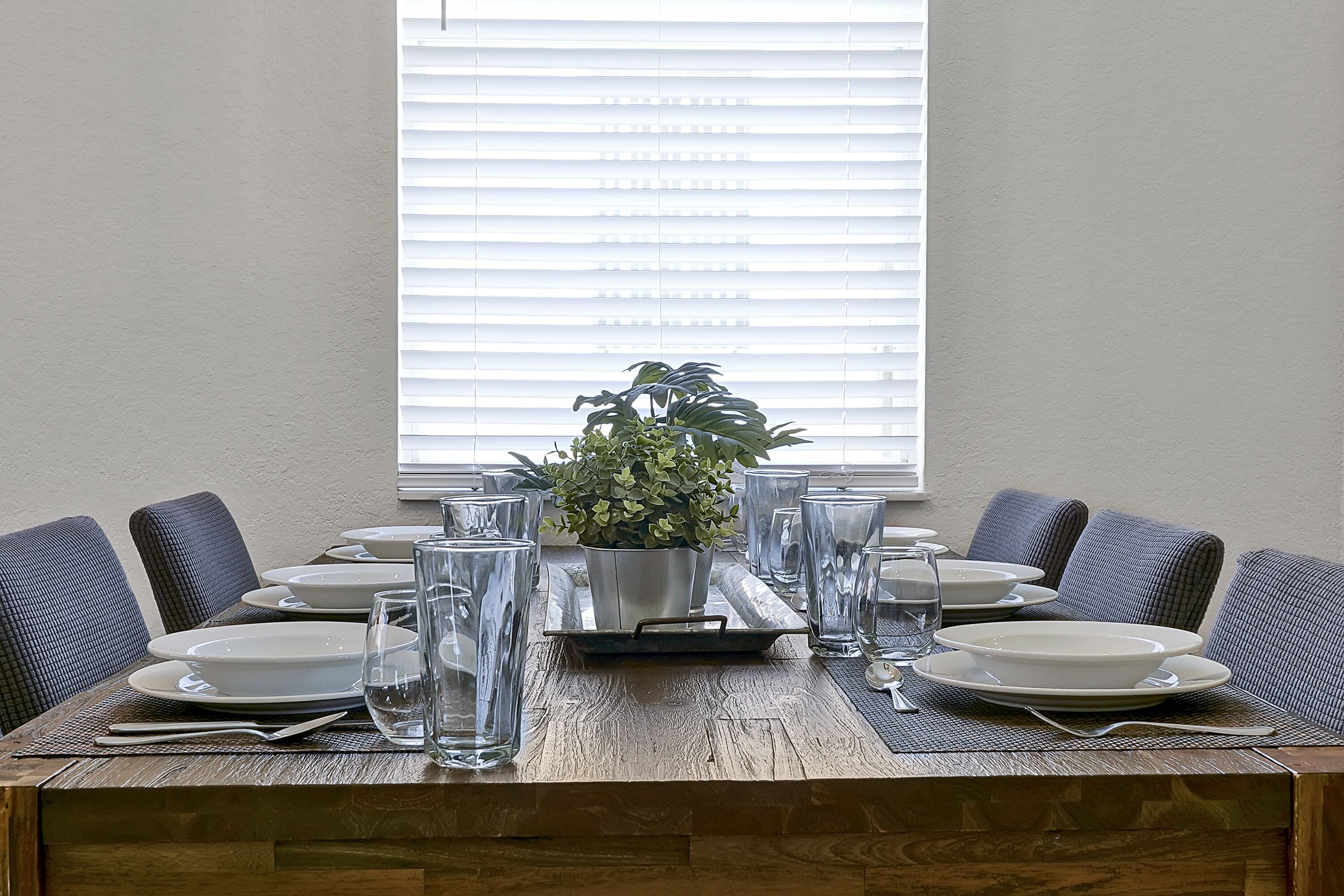 Una mesa de comedor dispuesta con platos, vasos y una planta en maceta, con una ventana al fondo.
