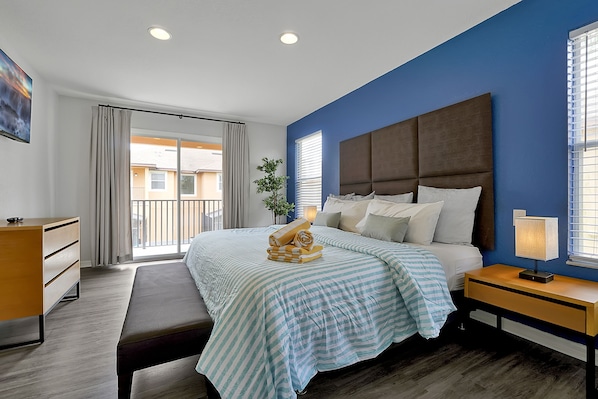 A modern bedroom with a large bed, a bedside table with a lamp, a bench, and a view of the outside.