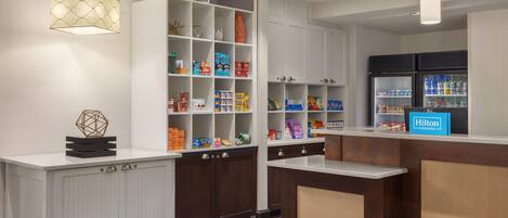 A hotel reception area with a counter, a welcome sign, and a variety of snack and beverage options on shelves.