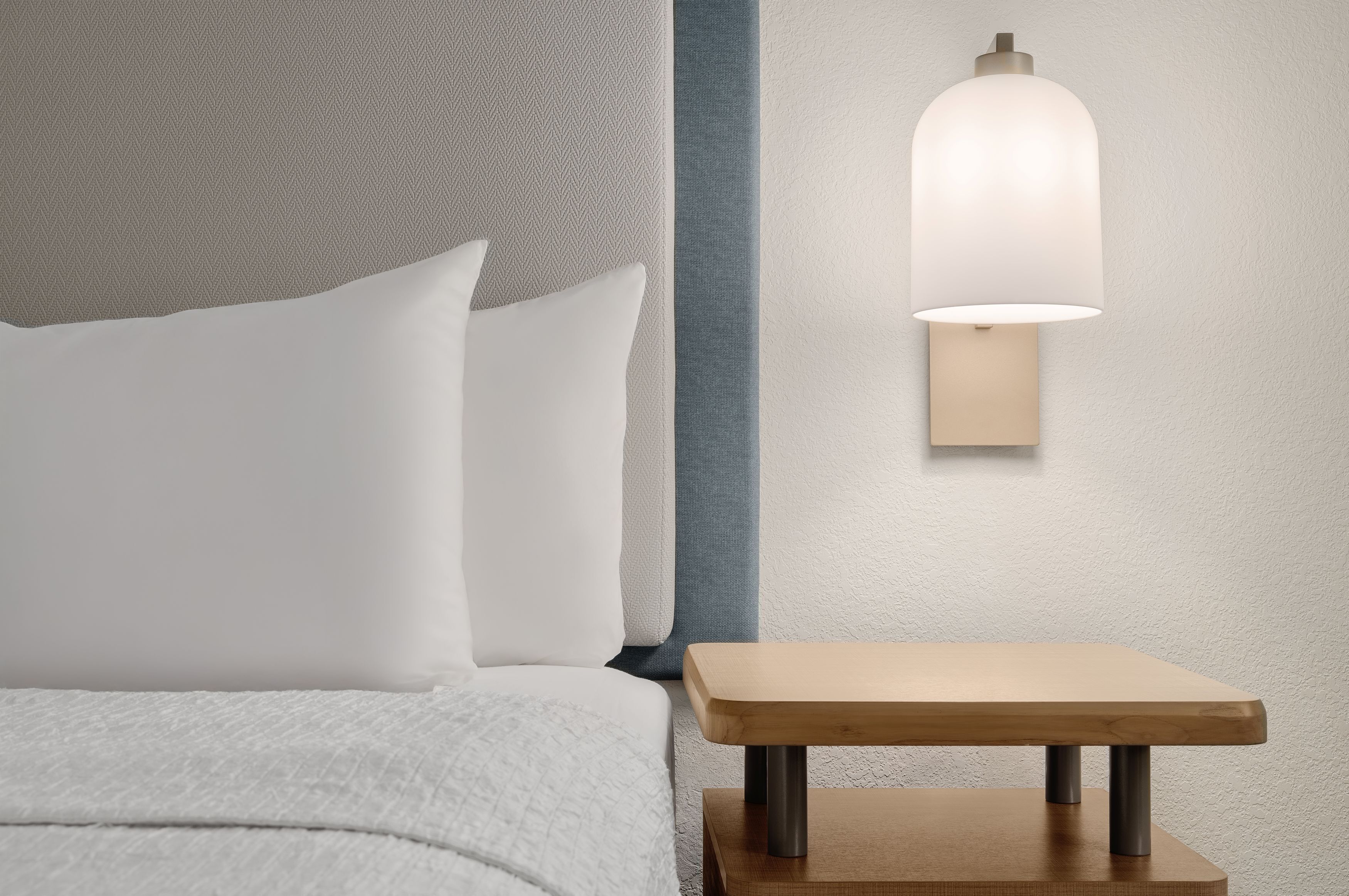 A neatly made bed with white bedding, a small wooden nightstand, and a wall-mounted light fixture.