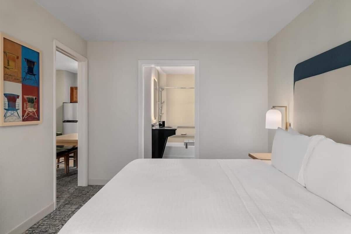 Quarto de hotel com uma cama grande, mesa de cabeceira, escrivaninha com cadeira, banheiro visível por uma porta aberta e um quadro na parede.