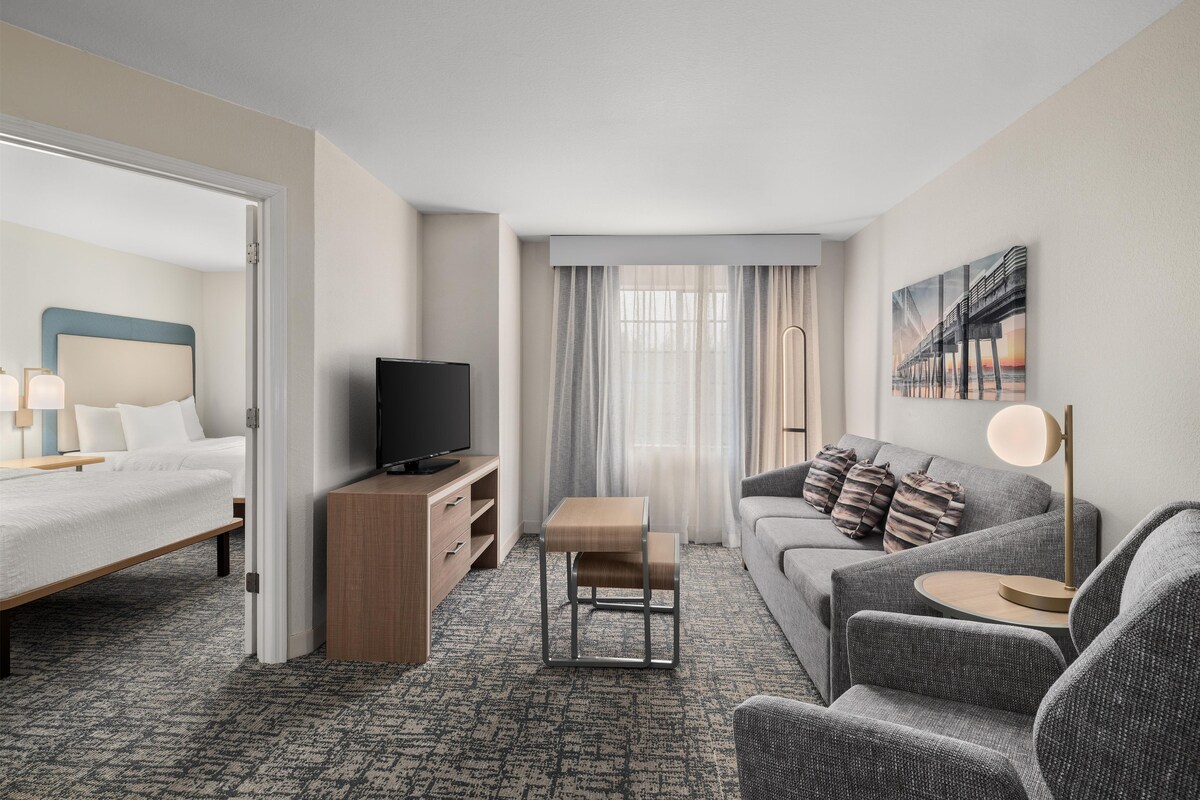 A hotel room with a bed, a TV, a sofa, a small table, and a painting on the wall.